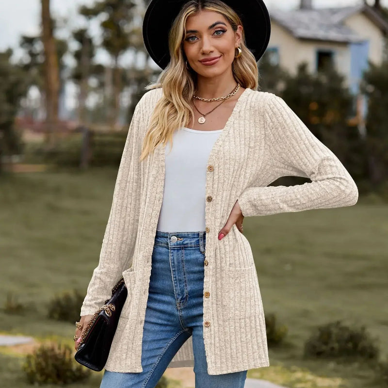Woman wearing a beige cardigan, white top, blue jeans, and black hat outdoors.
